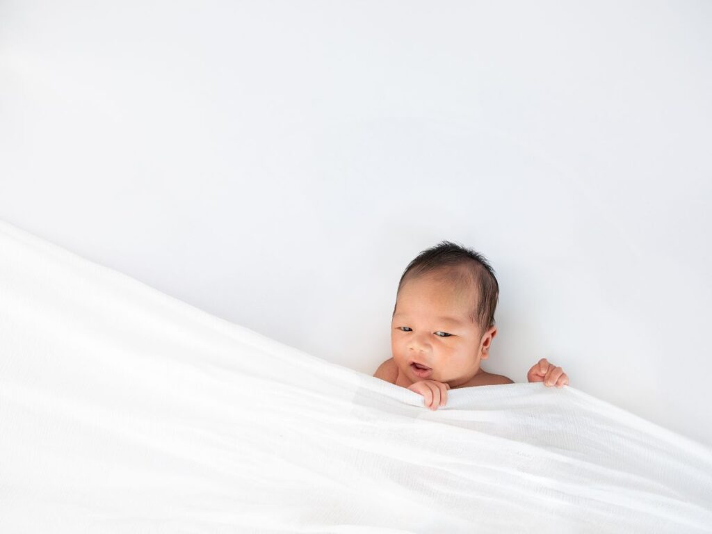 Curious newborn peeking out from a white blanket in a bright Pukekohe newborn photography session.