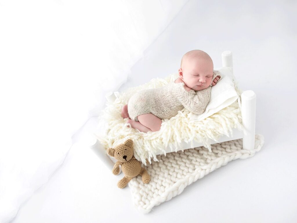 Sleeping newborn on a miniature bed with a teddy bear.