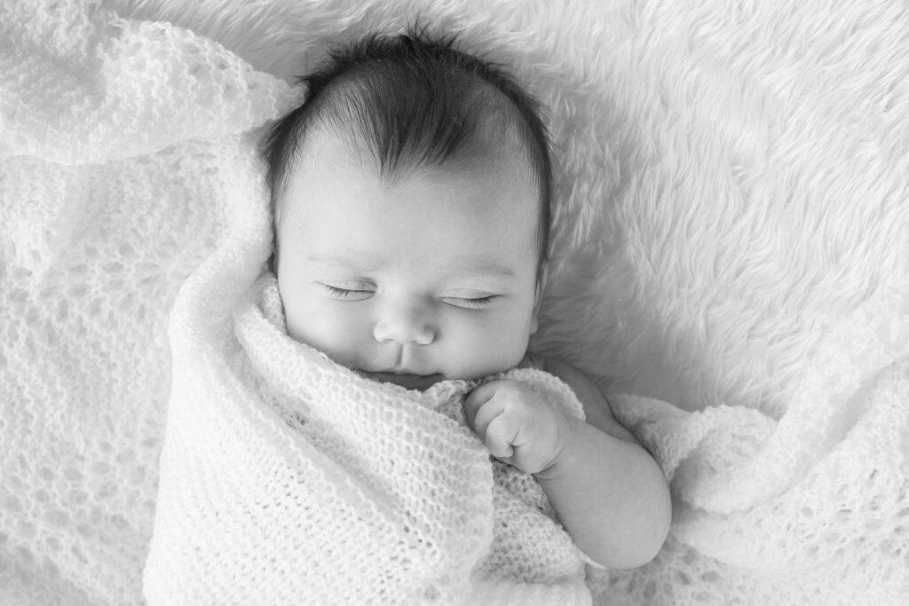 
A peaceful newborn wrapped in a delicate white blanket sleeps on a soft, fluffy surface in a black and white photograph.