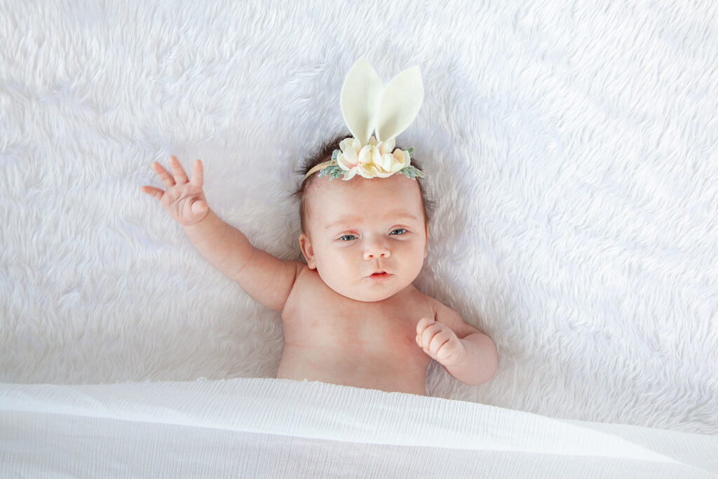 A newborn baby with a floral headband lies on a white fluffy blanket, gazing at the camera for a Pukekohe newborn shoot.