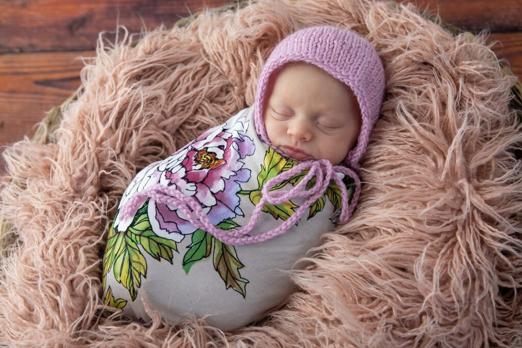 
A sleeping newborn is snugly wrapped in a floral swaddle and a purple knit hat, resting on a textured pink blanket with a rustic wood backdrop.
